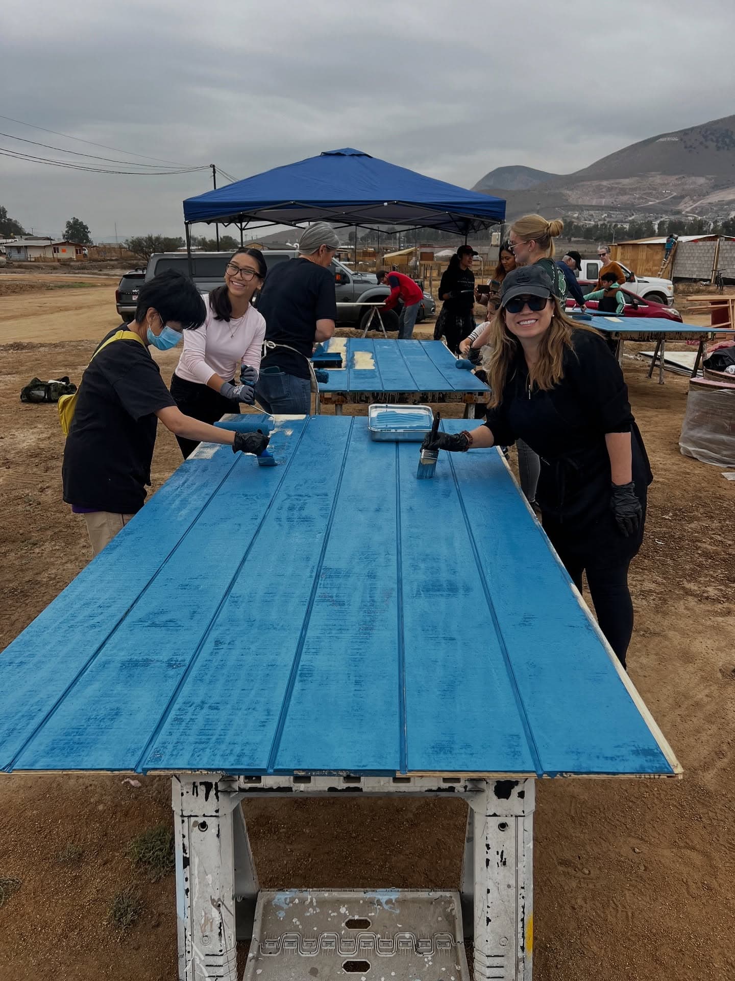 A long run of siding boards laid across sawhorses, being rolled with blue paint by volunteers under a canopy
